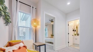 A modern living room with a sofa, a small table, and a floor lamp. There is a doorway leading to a bathroom with a mirror and a sink. - Walk 2 Beach | Game Room | Firepit |Heat/Salt Pool (St. Augustine)
