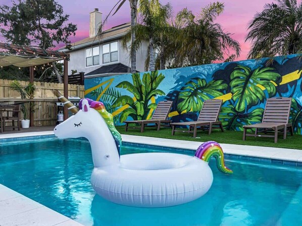 A swimming pool with a unicorn float, lounge chairs, and a mural of tropical leaves. - Walk 2 Beach | Game Room | Firepit |Heat/Salt Pool (St. Augustine)