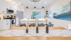 A modern kitchen with white cabinets, a central island, pendant lighting, and a large abstract painting on the wall. - Walk 2 Beach | Game Room | Firepit |Heat/Salt Pool (St. Augustine)