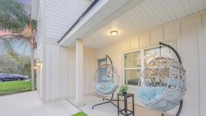 A covered porch with two wicker chairs and a small table, a white building, and a car parked outside. - Walk 2 Beach | Game Room | Firepit |Heat/Salt Pool (St. Augustine)
