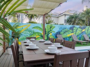 A dining area with a long table set for a meal, surrounded by chairs, with a mural of tropical foliage in the background. - Walk 2 Beach | Game Room | Firepit |Heat/Salt Pool (St. Augustine)