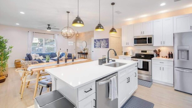 A modern kitchen with white cabinets, a central island, stainless steel appliances, pendant lighting, and a dining area with a wooden table and chairs. - Walk 2 Beach | Game Room | Firepit |Heat/Salt Pool (St. Augustine)