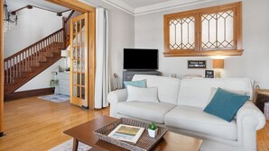 Una sala de estar con un sofá blanco, una mesa de centro con una bandeja y una planta, un televisor y una escalera con barandilla de madera.