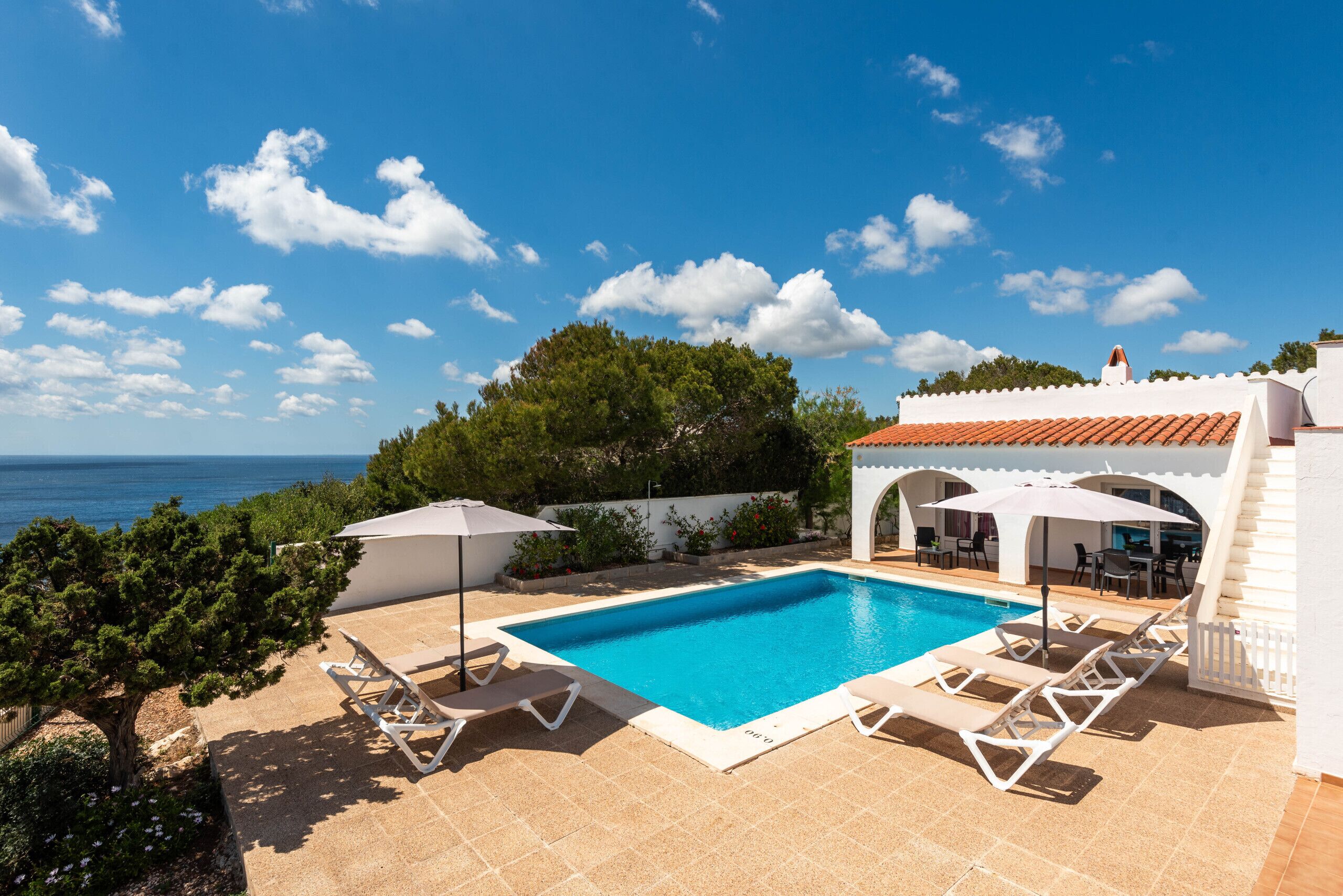 Une piscine entourée de chaises longues et de parasols, adossée à un bâtiment blanc doté d’un toit recouvert de tuiles rouges, et offrant une vue sur une étendue d’eau.