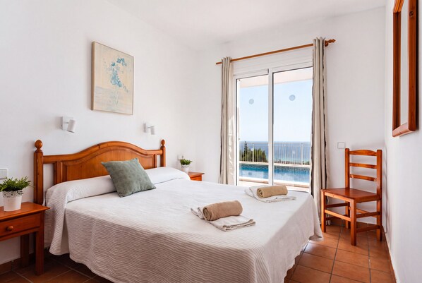 A neatly made bed with a headboard, a bedside table with a plant, a chair, and a window with curtains.