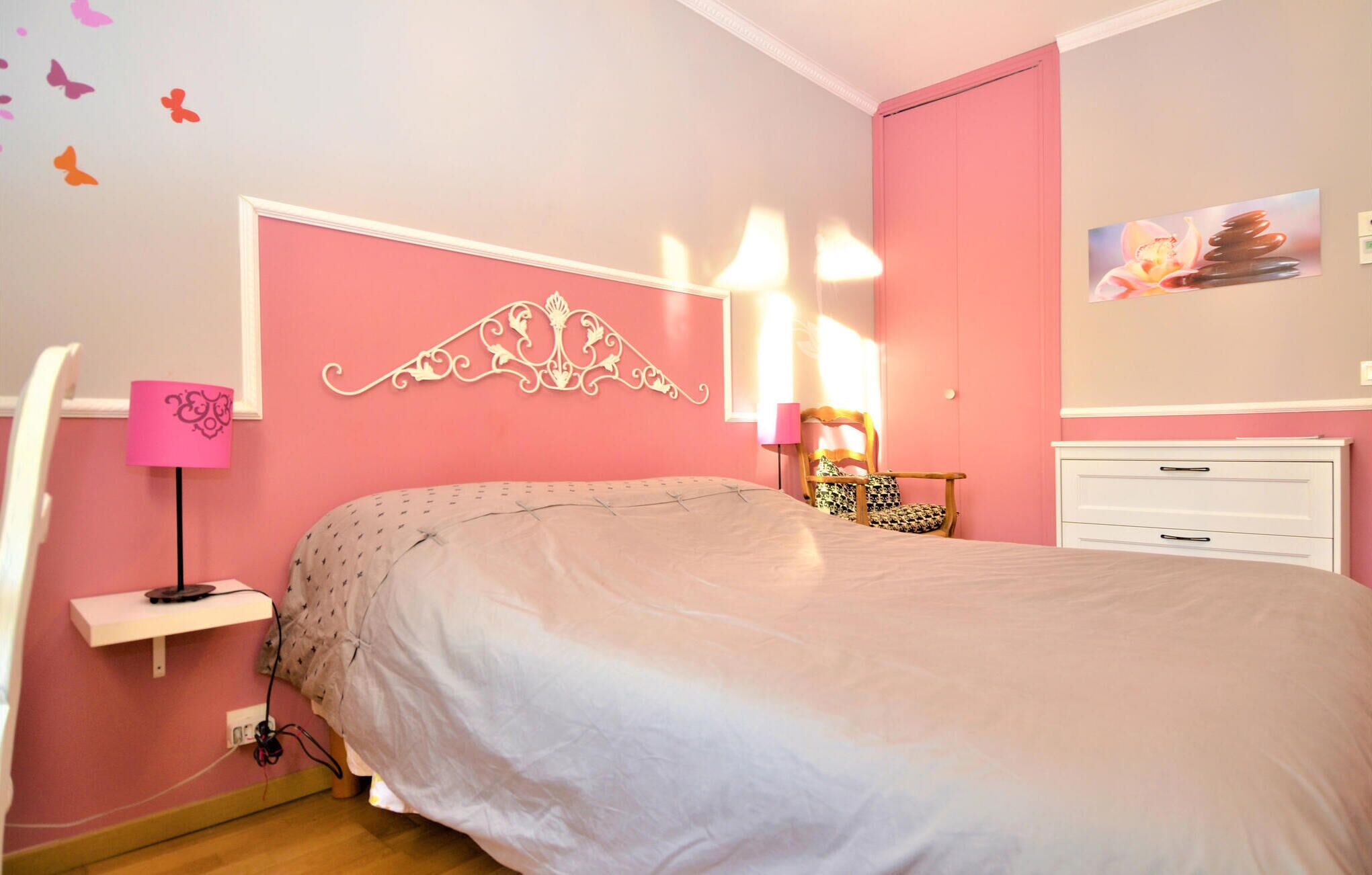 Een slaapkamer met een roze muur, een wit bed, een nachtkastje met een lamp, een stoel en een dressoir.