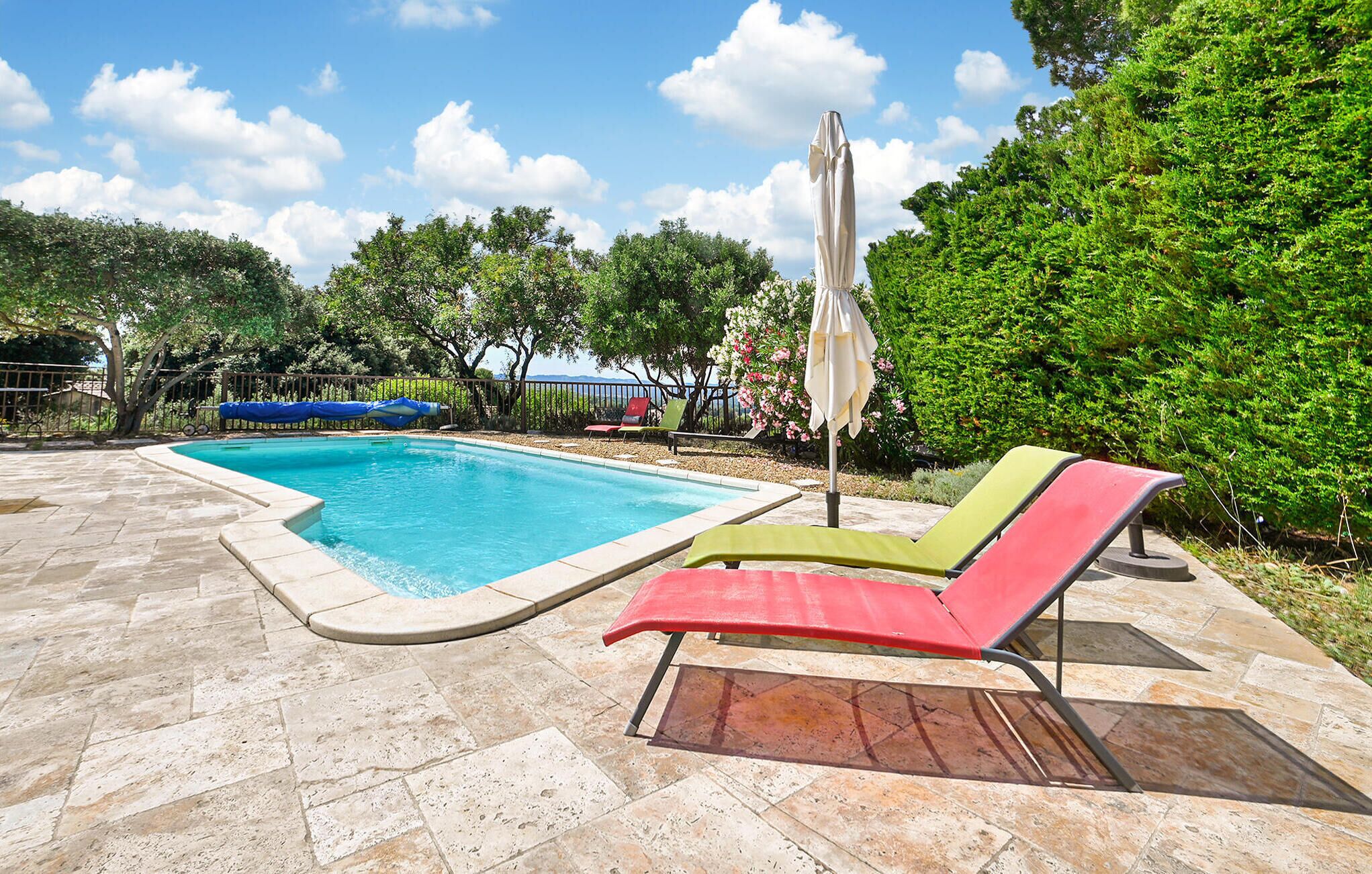 A swimming pool with a tiled deck, surrounded by lush greenery and a clear sky.