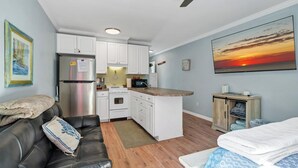 A compact living space with a kitchenette, a sofa, a bed, and a painting of a sunset.