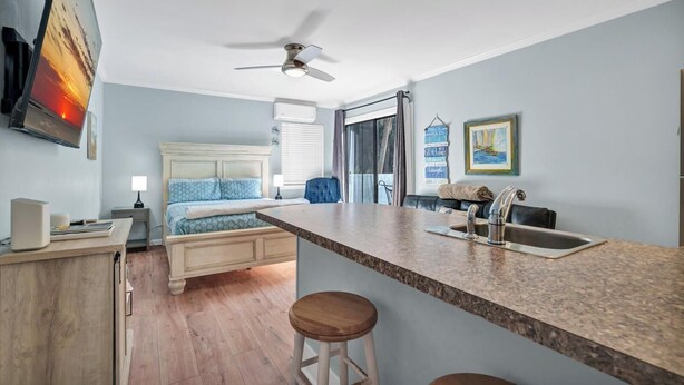 A compact hotel room with a bed, a desk, a chair, a small kitchenette, and a ceiling fan.