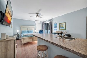Habitación de hotel pequeña con cama, escritorio, silla, kitchenette y ventilador de techo.