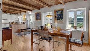 Eine geräumige Küche mit Holzbalken, ein Essbereich mit Tisch und Stühlen und große Fenster mit Blick ins Freie.