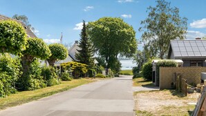 Eine ruhige Wohnstraße mit Häusern, einem großen Baum und einem Gewässer in der Ferne.