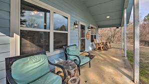 A covered porch with two chairs and a table, large windows, and a view of trees. - Hogden Home Near the Black Fork River & Trails! (Hodgen)