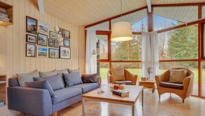 Un salon chaleureux avec un canapé gris, deux fauteuils en osier, une table basse en bois et une grande fenêtre agrémentée de rideaux.