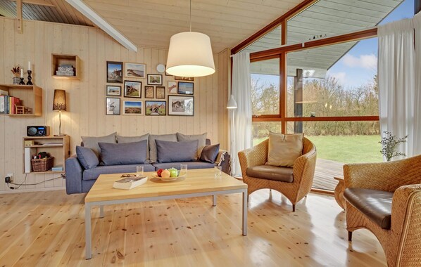 A cozy living room with a wooden floor, a blue sofa, a wicker armchair, and a coffee table. There are framed pictures on the wall and a large window with curtains. - Amazing home in Spøttrup with kitchen (Spøttrup)