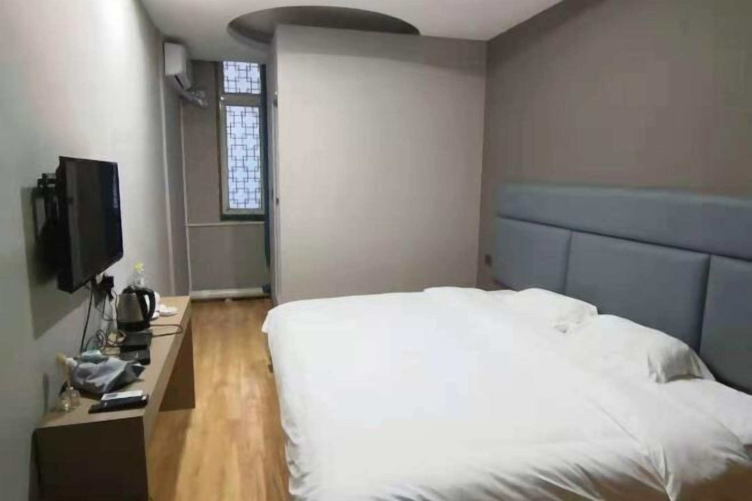 Un dormitorio con cama, televisor sobre un mueble, ventana y mesita de noche con tetera y taza.