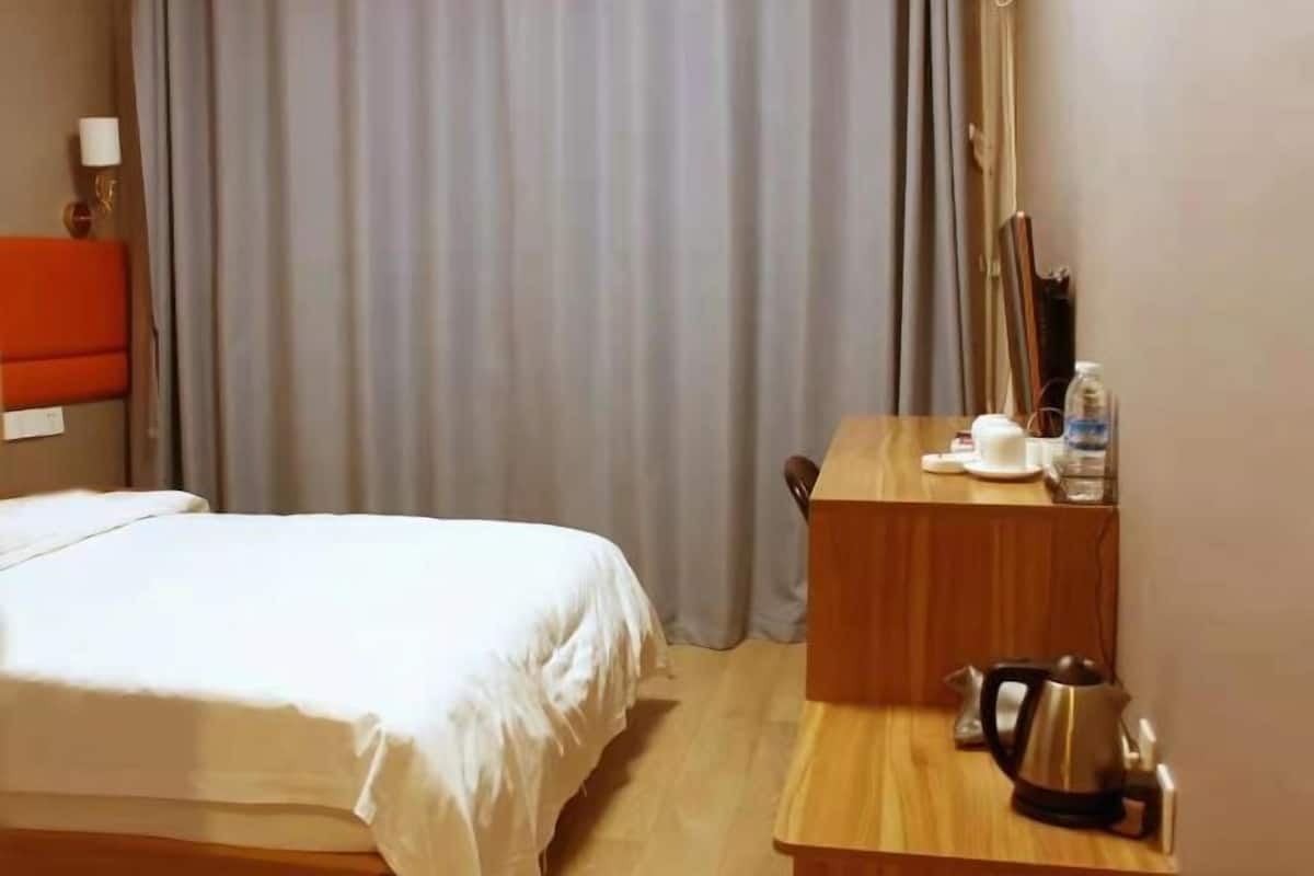 A hotel room with a bed, a wooden desk with a kettle, a cup, and a bottle of water.