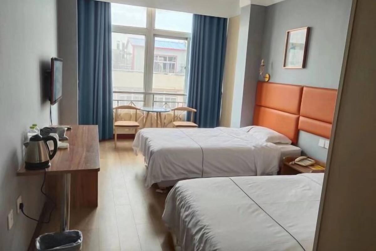 A hotel room with two beds, a desk with a kettle, a chair, a table, and a window with curtains.