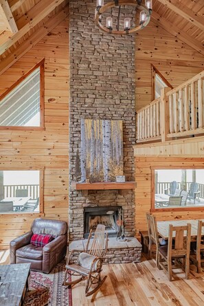 Interior de una cabaña de troncos con chimenea de piedra, mobiliario de madera y un balcón.