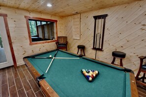 Een pooltafel met een groene viltbekleding, houten omlijsting en een raam met uitzicht op bomen.
