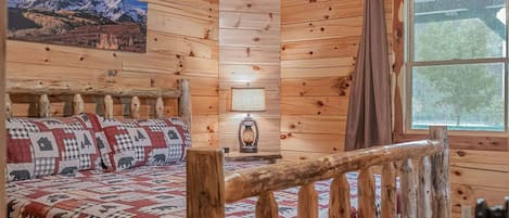 A wooden cabin interior with a bed, a ceiling fan, a window, and a wall-mounted picture.