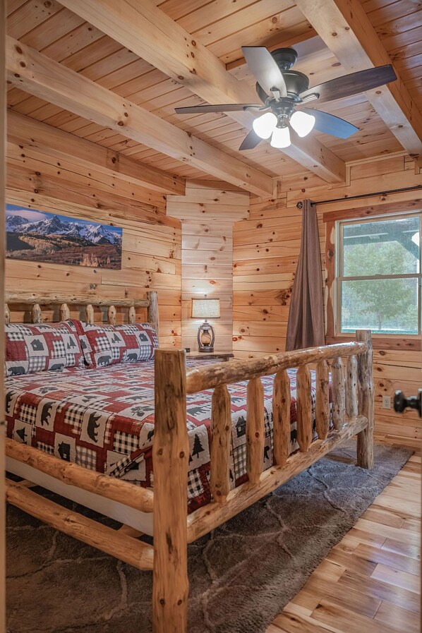 L’intérieur d’une cabane en bois, avec un lit, un ventilateur de plafond, une fenêtre et un tableau accroché au mur.