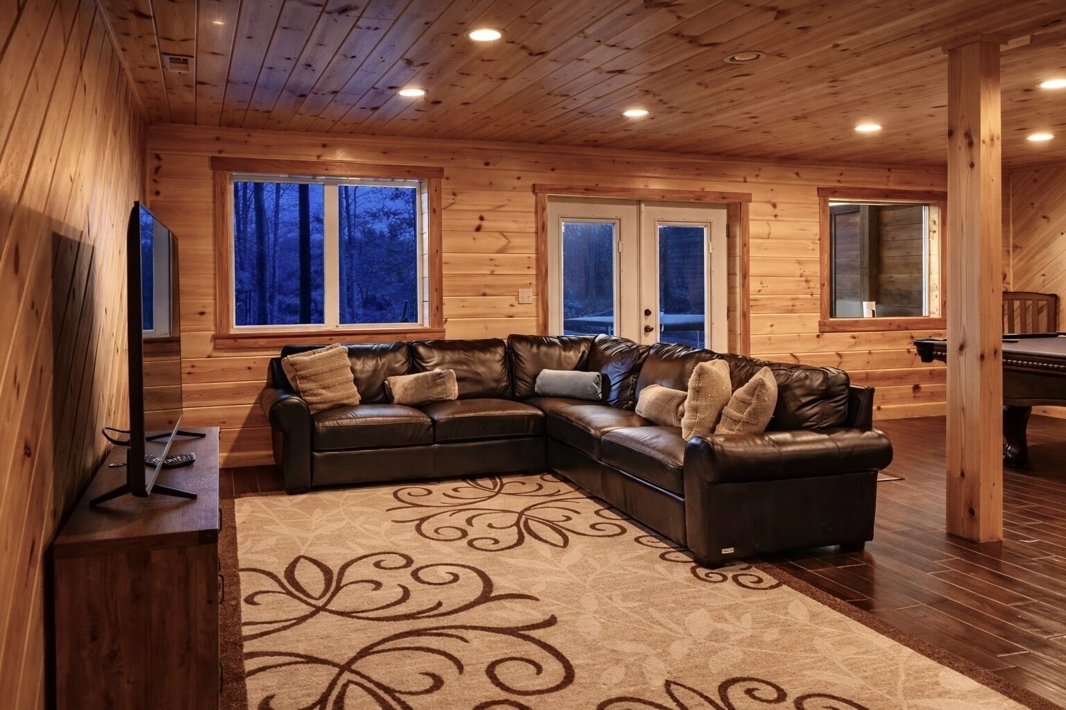A cozy wooden cabin interior with a leather sectional sofa, a large rug, and a pool table.
