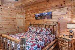 A wooden cabin room with a bed, a wooden headboard, a mounted picture of a mountain landscape, and a nightstand with a lamp.