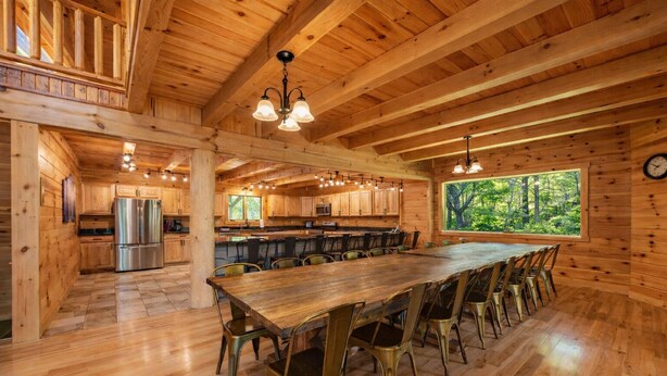 A spacious wooden cabin interior with a large dining table, chairs, a kitchen area, and a view of the forest through a large window. - Blue Ridge Lodge Hocking Hills: Accessible Luxury (Logan)