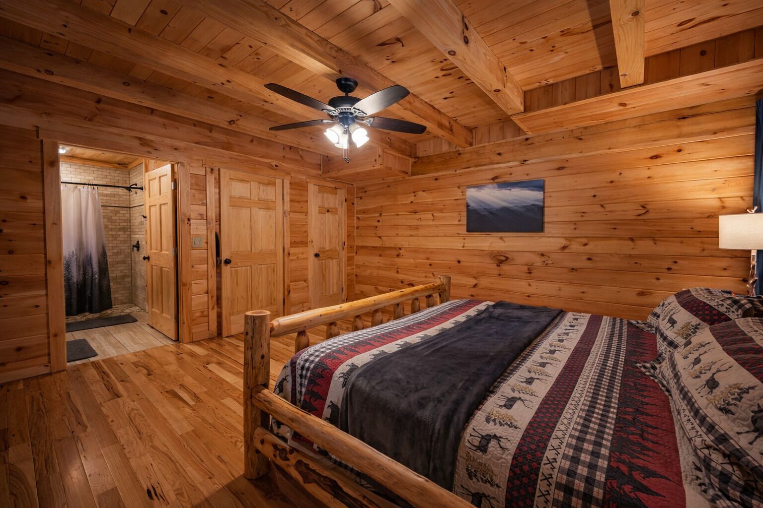 Interior de una cabaña de madera con una cama, un ventilador de techo y una puerta que da a un baño.