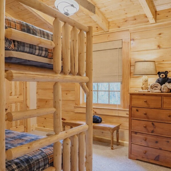 A wooden cabin interior with a bunk bed, a dresser, a lamp, and a window.