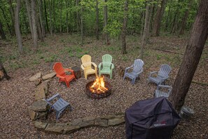 A campfire surrounded by Adirondack chairs in a forest setting.