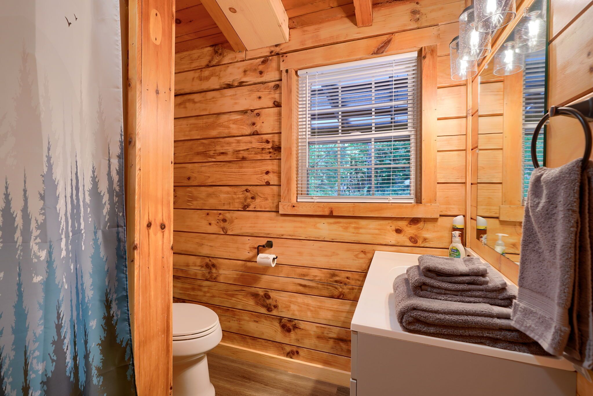 Un baño de cabaña de madera con un inodoro, una ventana con persianas, un lavamanos con una toalla y un espejo.