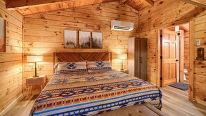 Ein Schlafzimmer in einer Holzhütte mit einem Bett, zwei Nachttischlampen, einem Nachttisch, einem Kleiderschrank und einer Tür zu einem weiteren Raum.