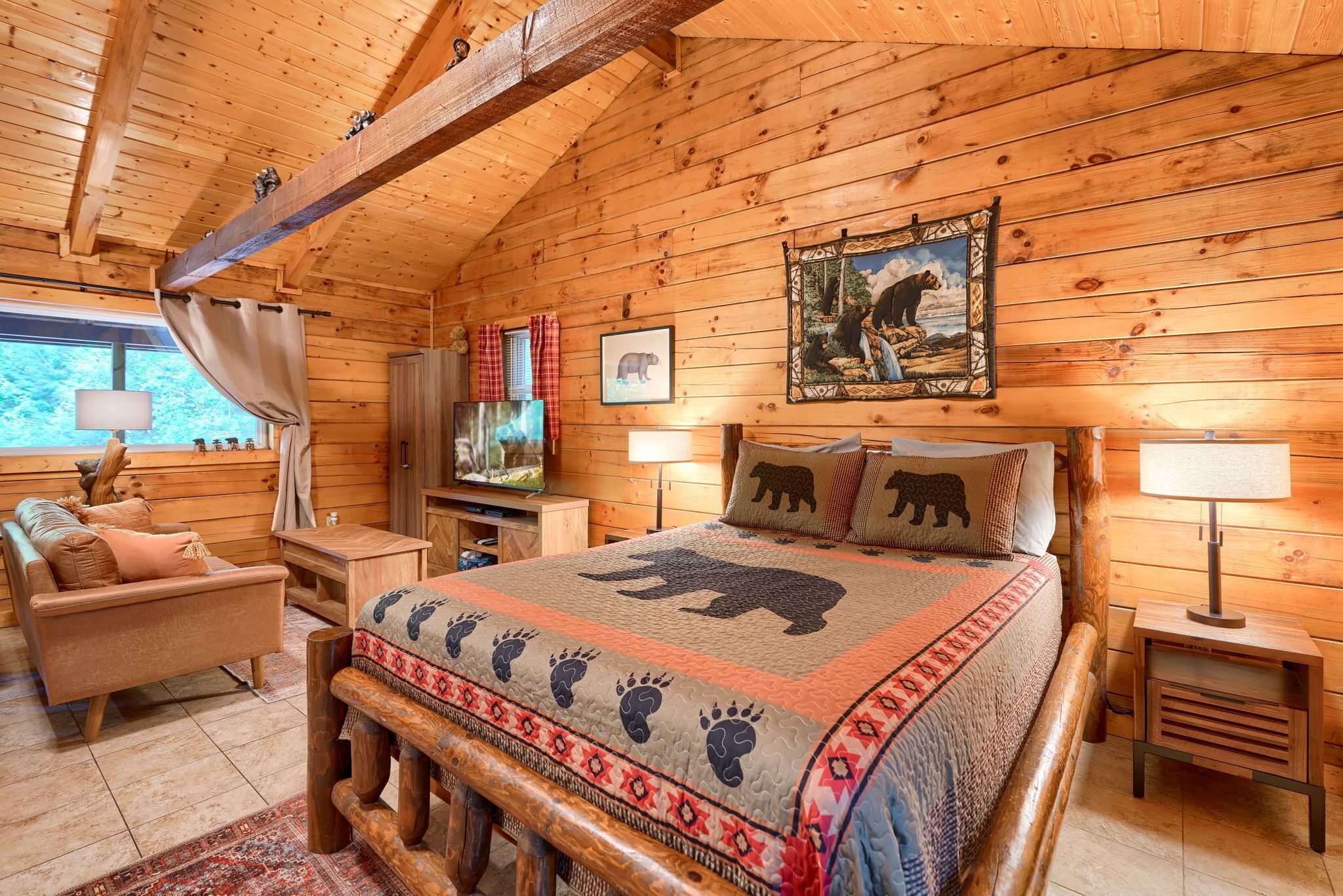 A log cabin interior with a bed, a sofa, a television, and a window with curtains.