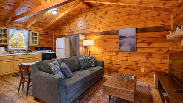 A cozy cabin interior with a wooden ceiling, a sofa, a dining table, and a kitchen area.