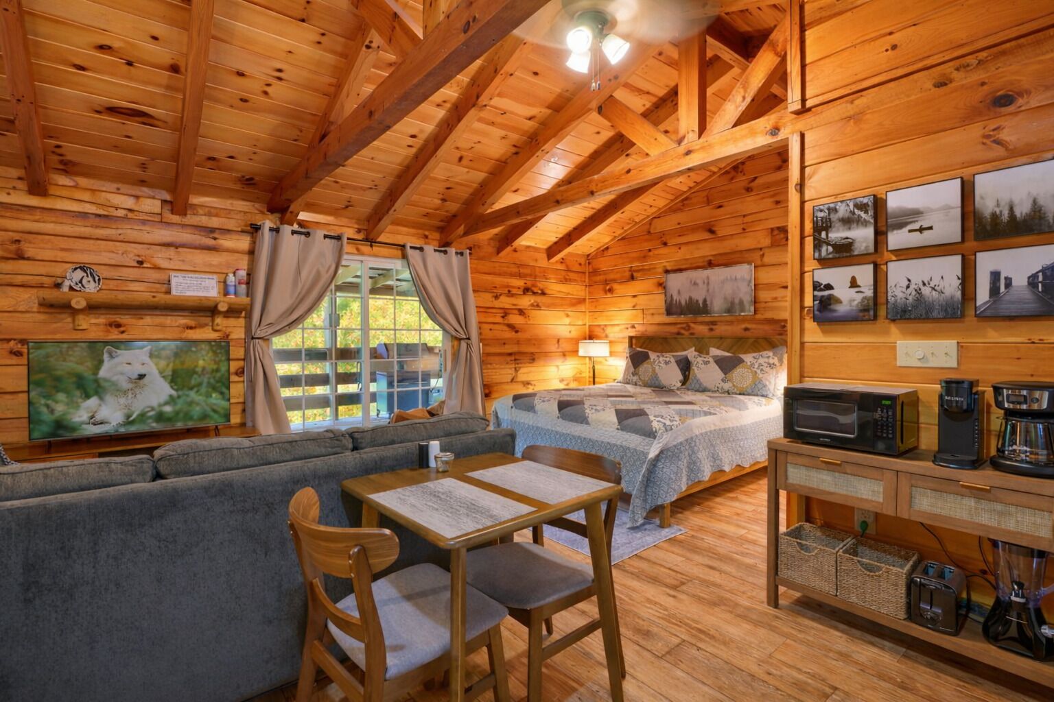 Interior acogedor de una cabaña con techo de madera, una cama con colcha, un sofá, una mesa de comedor con sillas y una cocina con electrodomésticos.