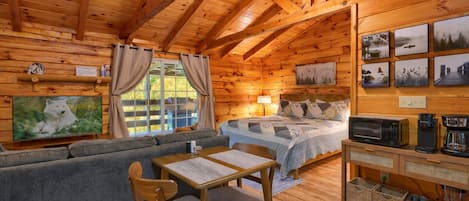 L’intérieur chaleureux d’un chalet avec un plafond en bois, un lit recouvert d’une courtepointe, un canapé, une table à manger avec des chaises et un coin cuisine équipé d’appareils électroménagers.