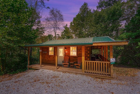 A small wooden cabin with a porch, a covered seating area, and a gravel driveway.