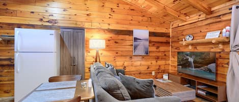 A cozy cabin interior with a wooden ceiling, a dining area with a table and chairs, a sofa, a television, and a refrigerator.