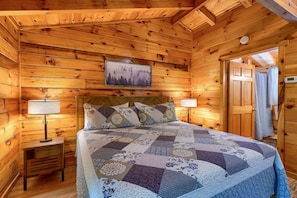 Un dormitorio de cabaña de madera con una cama, dos lámparas de noche, una mesita de noche, un cabecero, un cuadro colgado en la pared y una puerta con cortina de ducha.