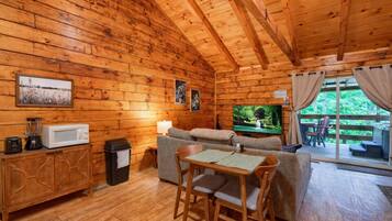 Un interior cálido de cabaña con detalles de madera, un sofá, un televisor, una mesa de comedor con sillas y vistas a través de un ventanal.