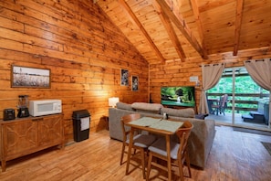 Un interior cálido de cabaña con detalles de madera, un sofá, un televisor, una mesa de comedor con sillas y vistas a través de un ventanal.