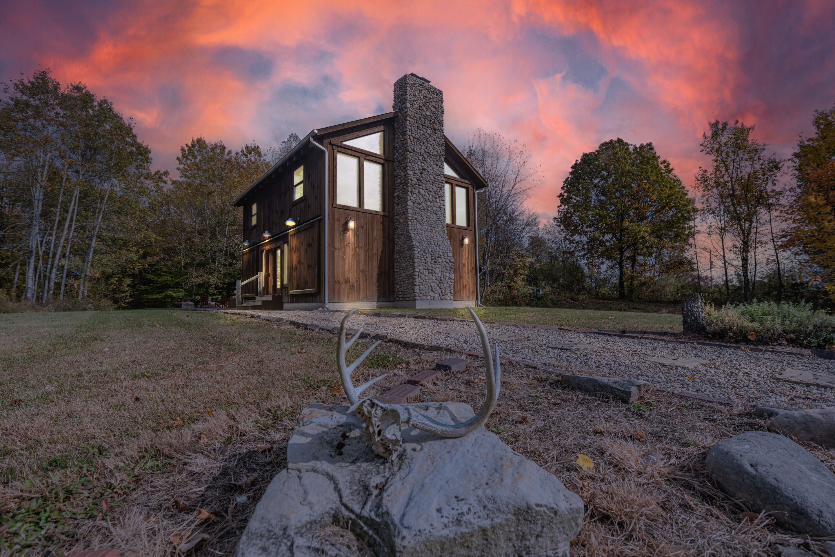 Una cabaña rústica con chimenea de piedra, puertas de madera y un camino de grava que conduce a una escultura de metal.