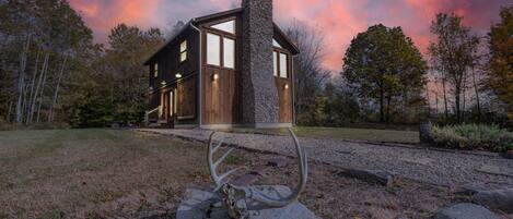 Een rustieke chalet met een stenen schoorsteen, houten deuren en een grindpad dat leidt naar een metalen sculptuur.
