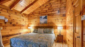Un dormitorio de cabaña de madera con una cama, dos lámparas de noche, una mesita de noche y un cuadro enmarcado en la pared.