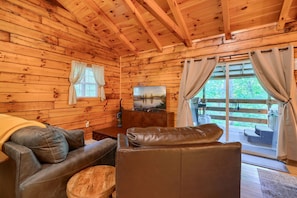 Interior acogedor de una cabaña con paredes de madera, un sofá de cuero, un televisor y un gran ventanal corredizo que da a una terraza exterior.