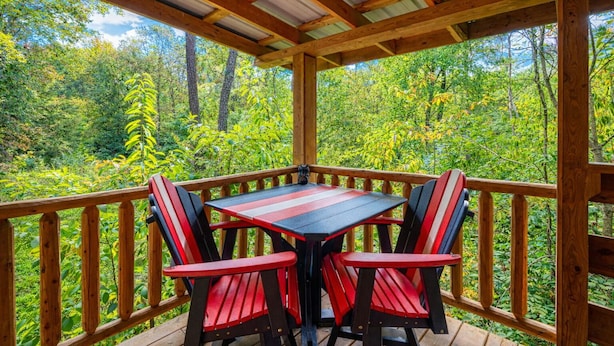 A covered outdoor area with a table and chairs, surrounded by a wooden railing, overlooking a forested landscape.