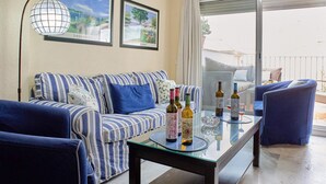 En stue med en stribet sofa, et glasbord med vinflasker og glas, en blå lænestol og udsigt gennem en skydedør.