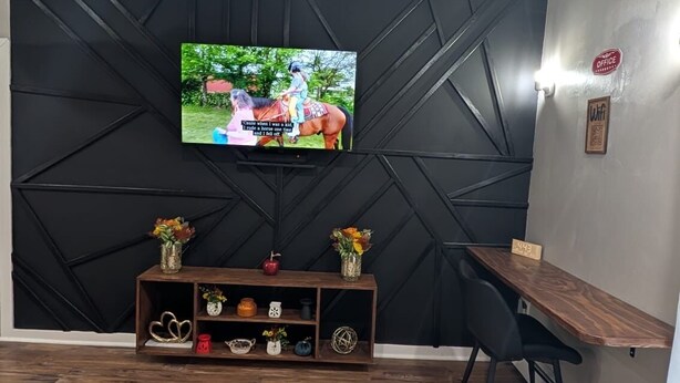 Un saló modern amb un televisió de pantalla plana penjada a una paret negra geomètrica, una taula baixa de fusta, una cadira negra i una catifa amb dibuixos.